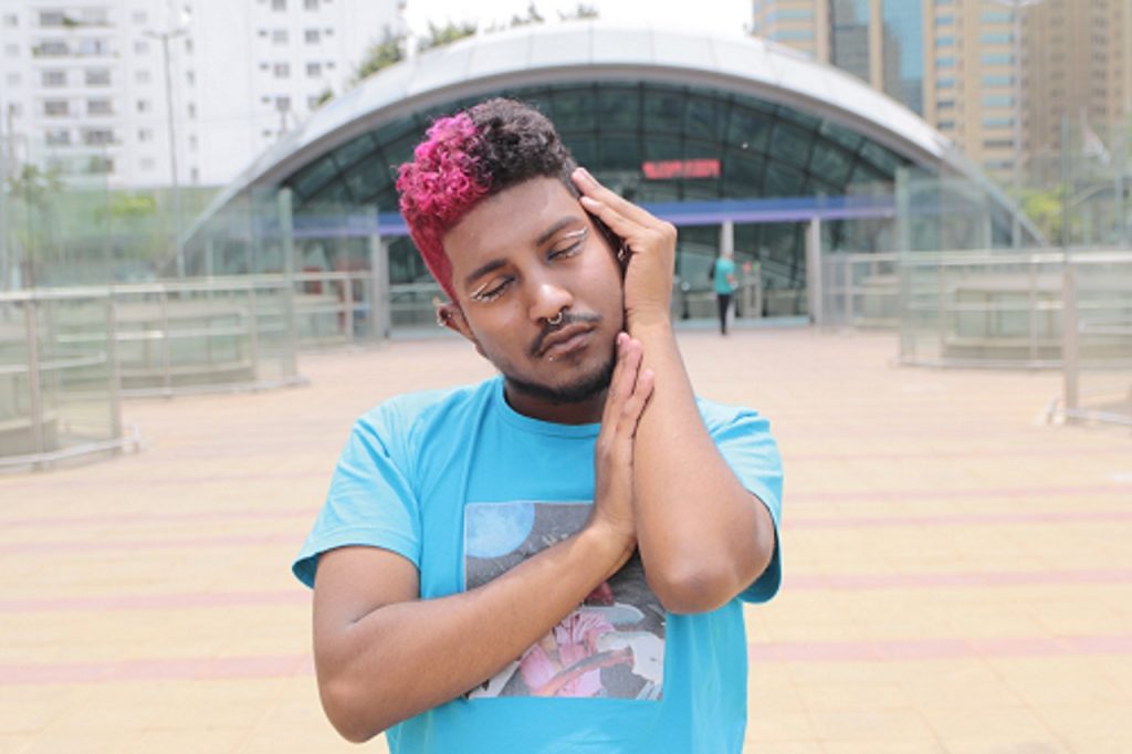 A picture of a Black person wearing a light blue t-shirt. Their hair is split-dyed black and pink and have a septum nose piercing as well as graphic white eyeliner.
